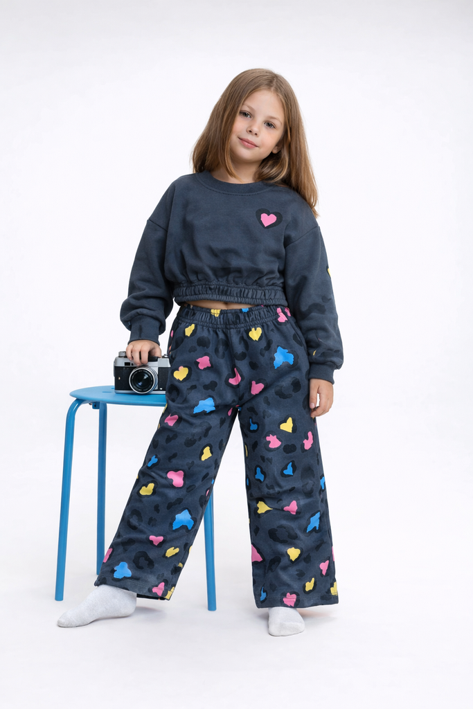 Young girl wearing a dark blue outfit with colorful heart patterns, standing next to a small table with a camera on it.